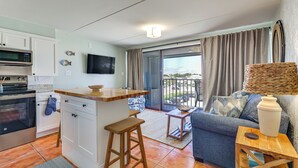A compact kitchen with a microwave, oven, and a small island with stools. A living area with a sofa and a coffee table is visible, featuring a large window and a balcony. - Carolina Beach Condo w/ Marina View, on Boardwalk (Carolina Beach)