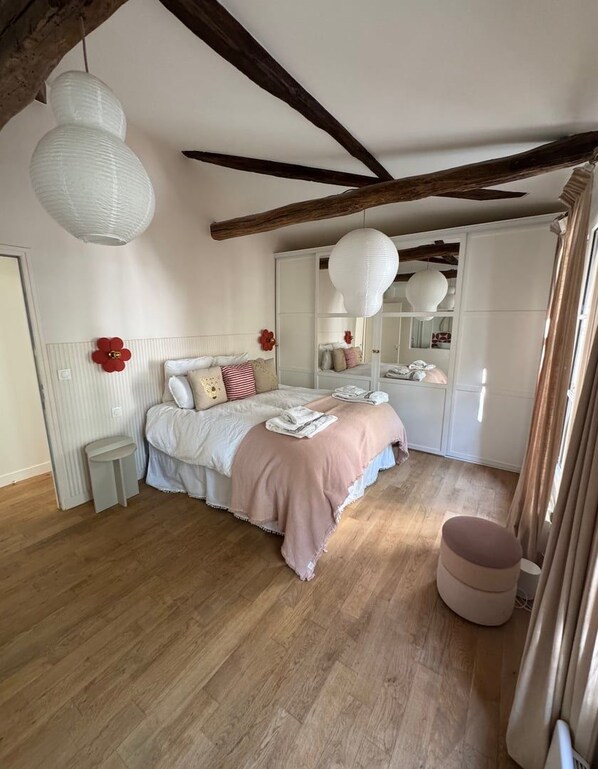 A bedroom with a wooden floor, a bed with a pink blanket, a small bedside table, a mirror, and a wardrobe.