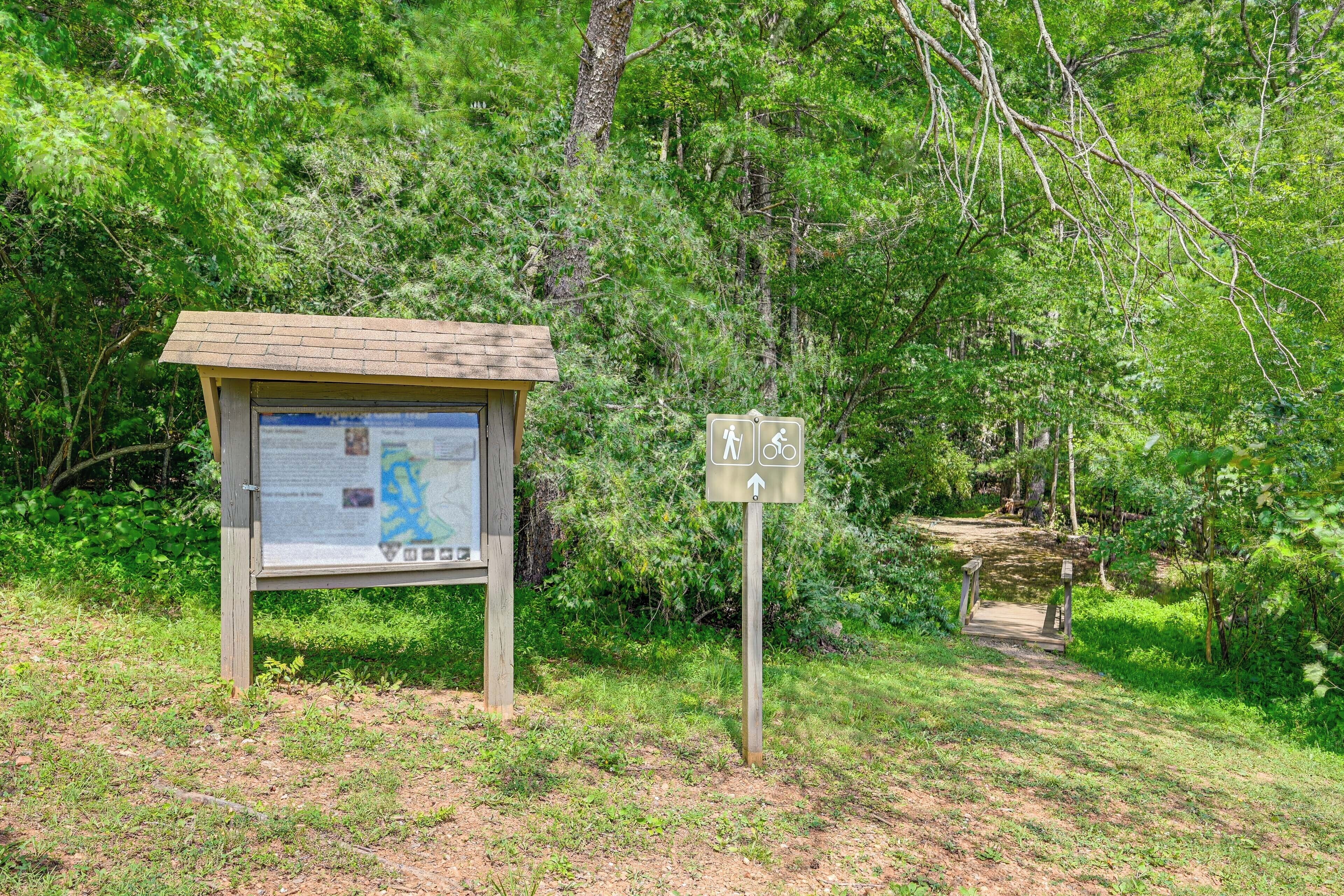 Ein hölzernes Wegweiser mit Wanderschildern und Radwegsymbolen, eine kleine Schutzhütte mit Anschlagtafel und ein Feldweg, der in einen Wald führt.