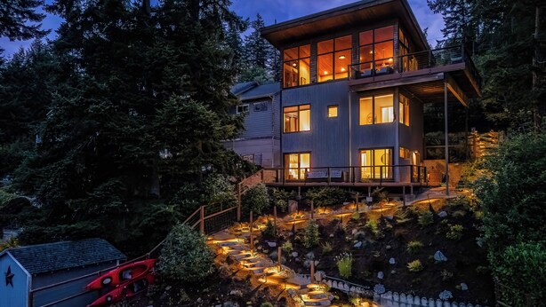 Une maison moderne à deux étages, dotée d’un balcon, entourée d’un jardin aménagé avec des marches en pierre et un foyer extérieur.