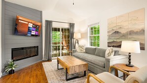 A modern living room with a flat-screen TV mounted on a grey wall, a fireplace, a grey sofa, a wooden coffee table, and a painting of mountains on the wall.