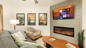 A living room with a sofa, armchair, coffee table, fireplace, mounted TV, and framed posters on the wall.