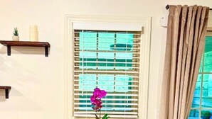 A dining area with a round table, two black chairs, a vase with a purple flower, a window with blinds, and a ceiling vent.