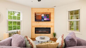 A cozy living room with a fireplace, a flat-screen TV mounted on a wooden wall, and a comfortable seating area with cushions. - Hot Tub · Fire Pit · EV Charger · Near Asheville (Asheville)