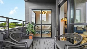 Un balcon doté de meubles en osier, d’une plante en pot et d’une porte-fenêtre coulissante donnant sur un espace de vie avec un canapé et une lampe.