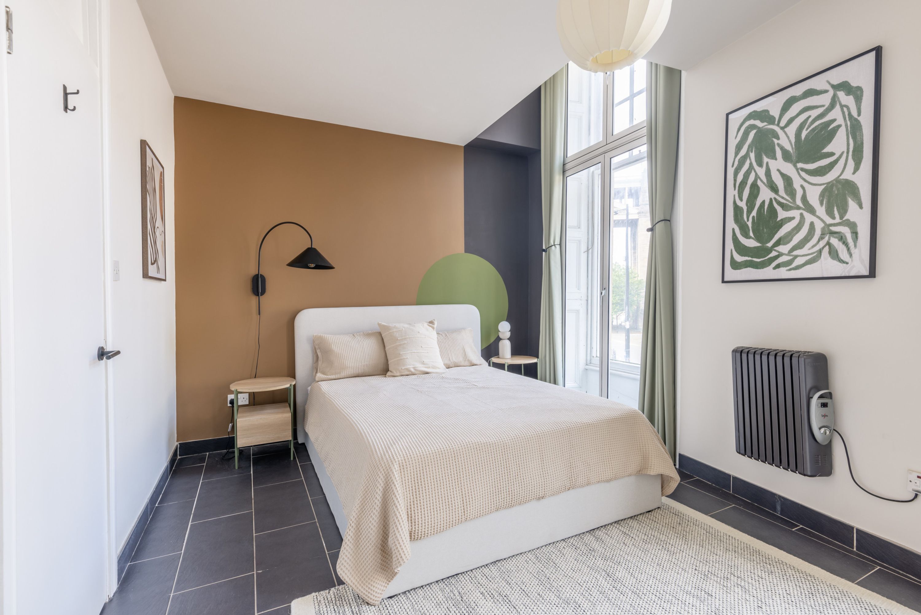 Een slaapkamer met een bed, nachtkastje, lamp, radiator en kunstwerk.
