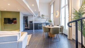 Ein modernes Wohnzimmer mit einer Couch, einer Essecke mit Tisch und Stühlen, einer Küche im Hintergrund und großen Fenstern, die für natürliches Licht sorgen.