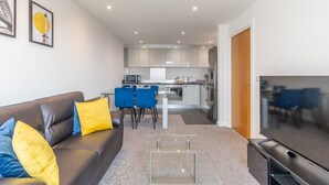 Un salon moderne avec un canapé en cuir marron, des coussins bleus et jaunes, une table basse en verre et un téléviseur à écran plat. Au fond, on aperçoit une cuisine avec des meubles blancs, un coin repas avec des chaises bleues et un grand miroir qui reflète la pièce.