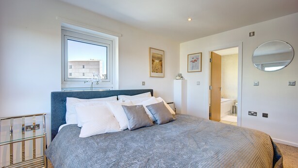 A modern bedroom with a large bed, a bedside table, a window, and a bathroom visible through an open door. - Atico in Newcastle Upon Tyne (Newcastle-upon-Tyne)