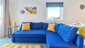 Ein modernes Wohnzimmer mit einem blauen Modulsessel, einem kleinen Beistelltisch, einer gelben Tagesdecke, einem großen abstrakten Gemälde, einem Fenster mit Blick und einem Essbereich mit gelben Stühlen.