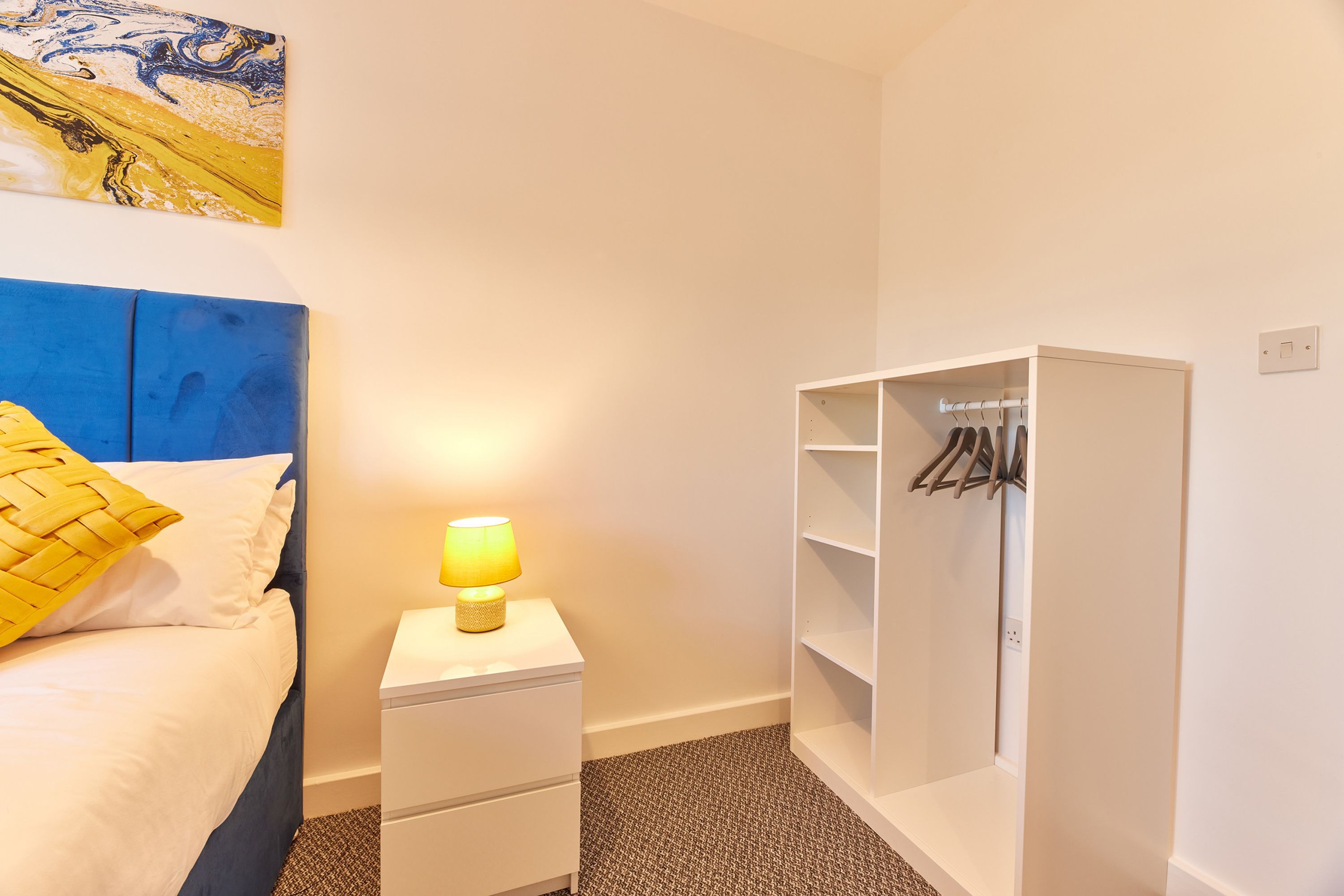 Ein Schlafzimmer mit Bett, Nachttisch mit Lampe, Kleiderschrank und einem Bild an der Wand.