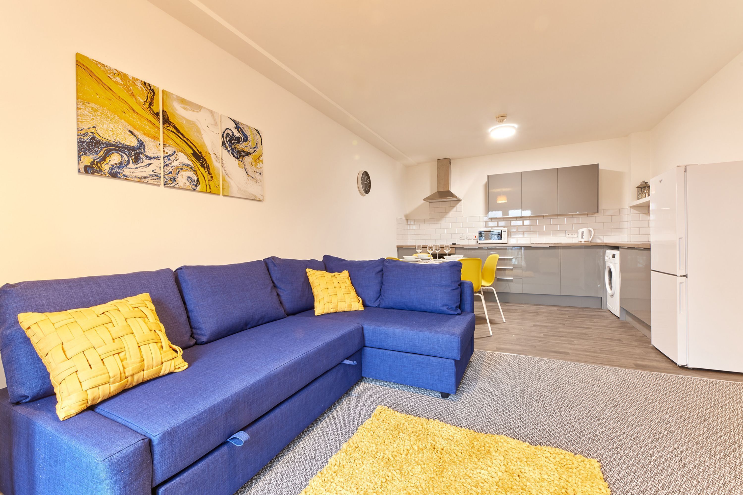 Een moderne woonkamer met een blauwe modulaire bank, een geel geweven sierkussen en een groot geel vloerkleed. In de achtergrond is een keuken met witte apparaten en een grijze achterwand zichtbaar.