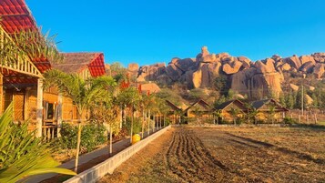 Un pueblo rural con casas de madera tradicionales, un camino de tierra y una imponente montaña rocosa al fondo.