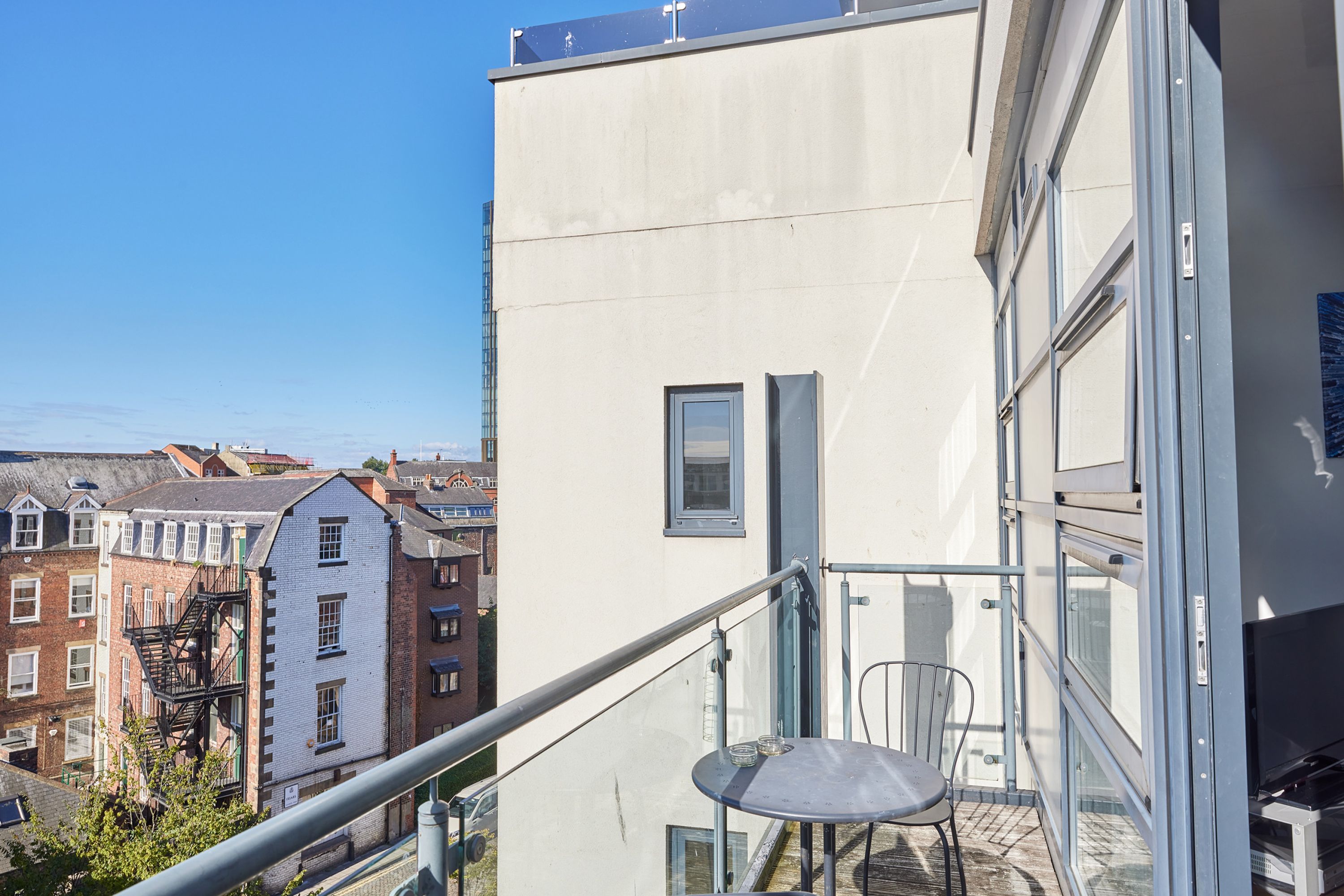 A balcony with a chair and a small table, overlooking a cityscape with various buildings.