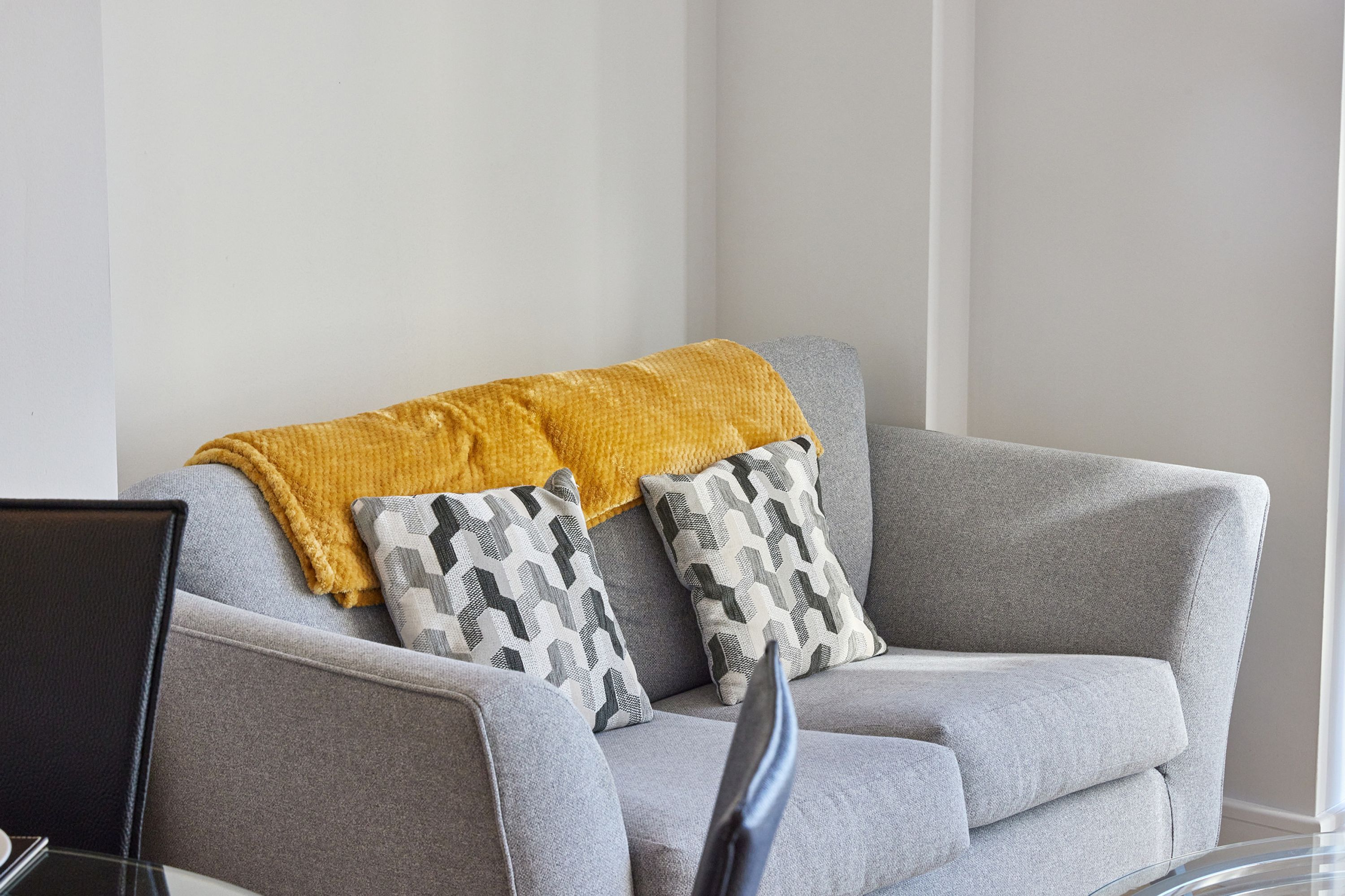 A grey sofa with a mustard throw blanket and patterned pillows, a black chair, and a glass table with a laptop.
