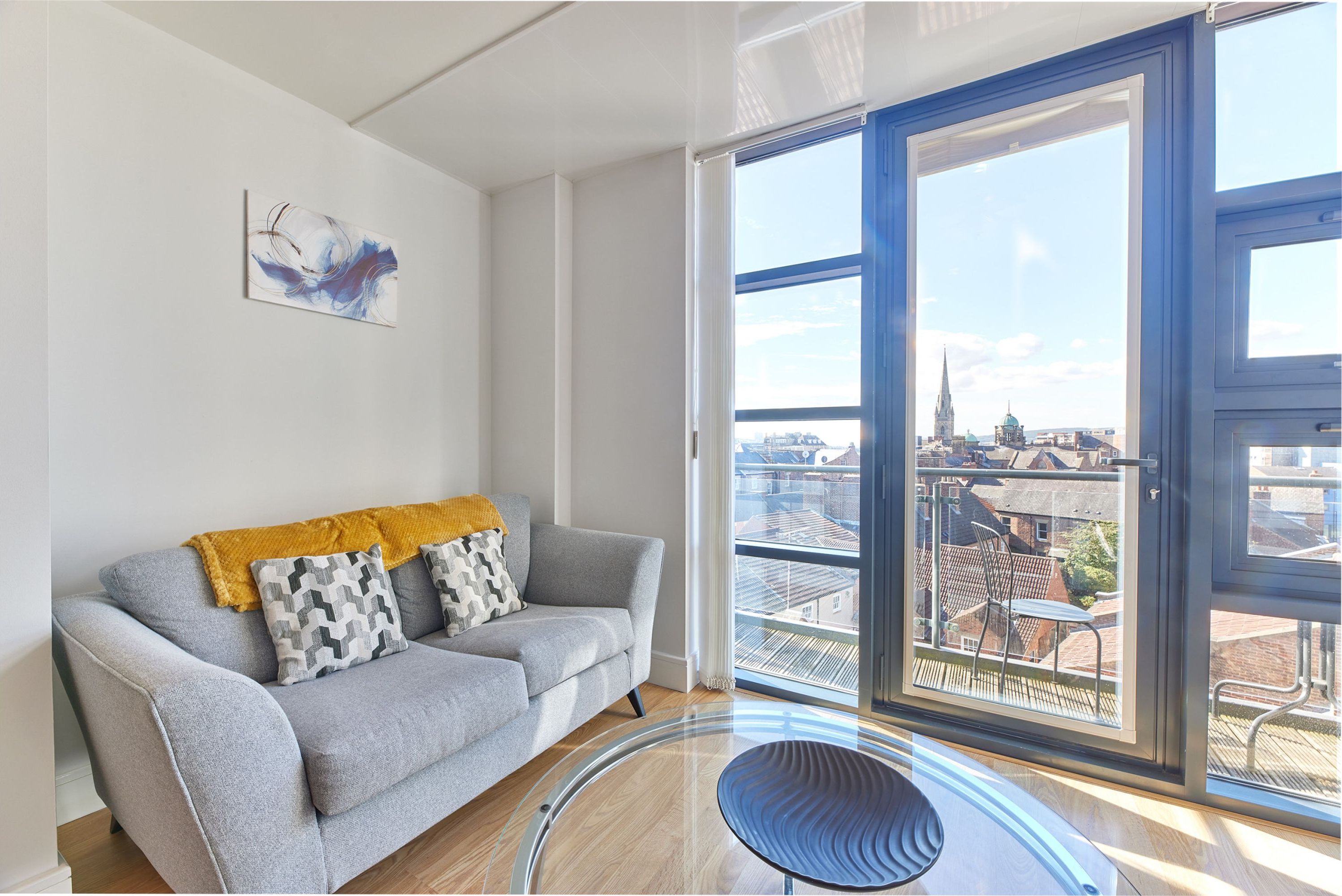 Ein modernes Wohnzimmer mit einem grauen Sofa, einer gelben Decke und einem schwarzen Sessel. Ein großes Fenster bietet Blick auf eine Stadtansicht.