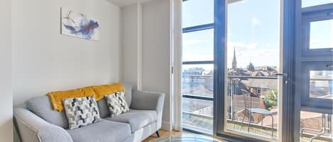 A modern living room with a grey sofa, a yellow throw blanket, and a black chair. A large window offers a view of a cityscape.