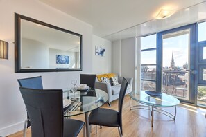 A modern living room with a glass dining table, black chairs, a grey sofa with yellow cushions, and a large window offering a city view. - Low Friars in Newcastle Upon Tyne (Newcastle-upon-Tyne)