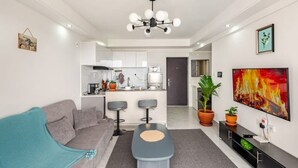 Ein modernes Wohnzimmer mit einem grauen Sofa, einem Couchtisch und einem Fernseher. Im Hintergrund befindet sich eine Küche mit weißen Schränken und ein Essbereich mit zwei Barhocker.