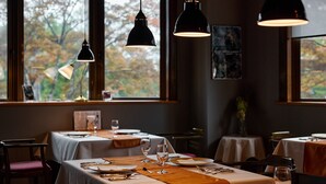 Un restaurant avec des tables dressées pour le dîner, de grandes fenêtres offrant une vue sur le feuillage automnal et des lampes suspendues décoratives.