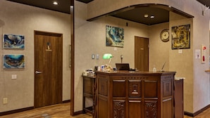 Un espace d’accueil d’hôtel avec un comptoir de réception en bois, des œuvres d’art encadrées accrochées aux murs et une horloge.