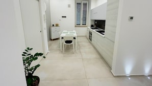 Een kleine keuken met witte kasten, een eettafel met stoelen, een raam met jaloezieën en een plant in pot.