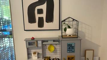 Une cuisine miniature pour enfants, avec un évier, un four et des tiroirs de rangement, un tableau abstrait encadré et une plante en pot posée sur une étagère.