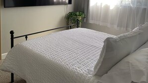 Ett sovrum med en säng, en tv som är monterad på väggen, ett fönster med tunna gardiner och en krukväxt.