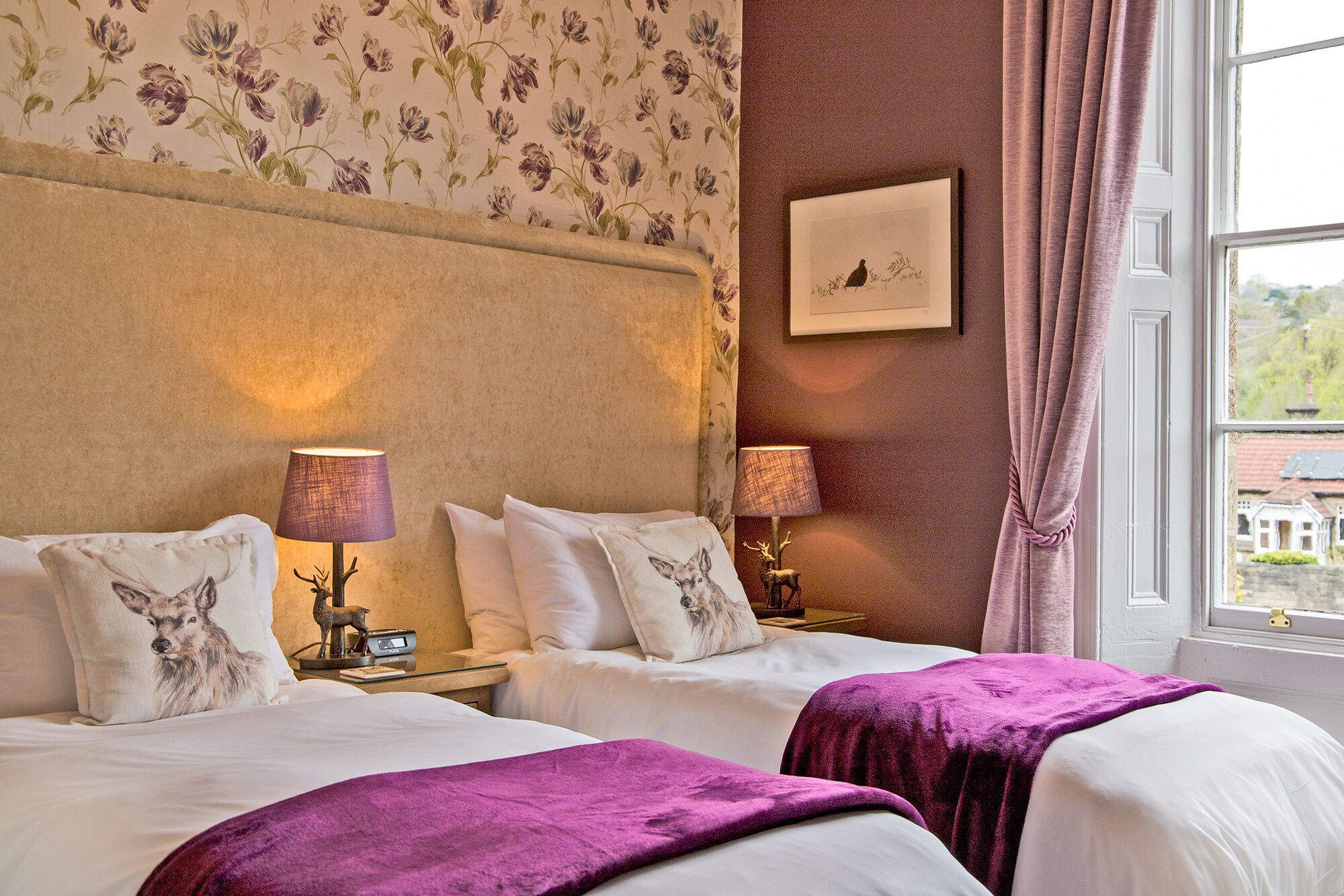 A hotel room with two beds, floral wallpaper, a framed picture on the wall, and a window with curtains.