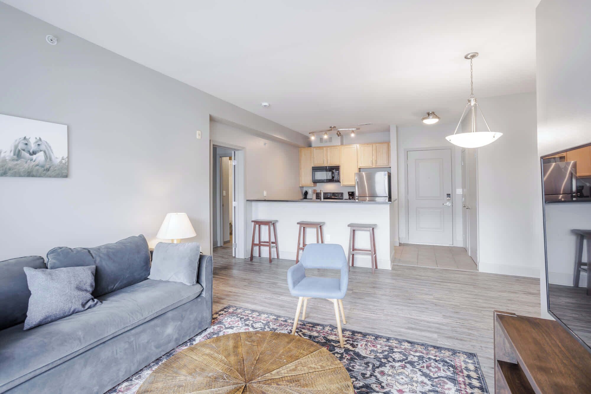 Ein modernes Wohnzimmer mit grauem Sofa, einem Couchtisch und einer Küchenzeile mit Holzausstattung und einem Edelstahlkühlschrank.