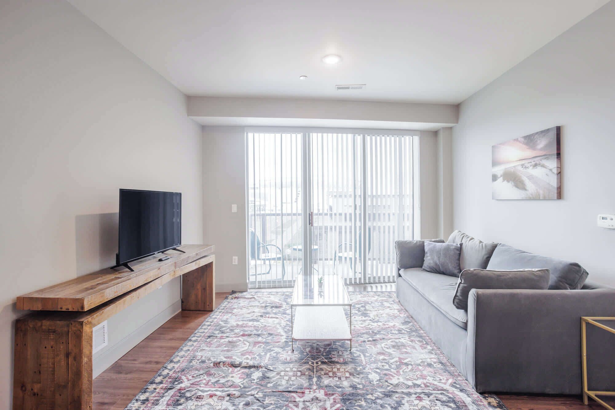 Ein modernes Wohnzimmer mit einem Flachbildfernseher auf einem Holzständer, einem grauen Sofa, einem gemusterten Teppich und einem großen Fenster mit Jalousien.