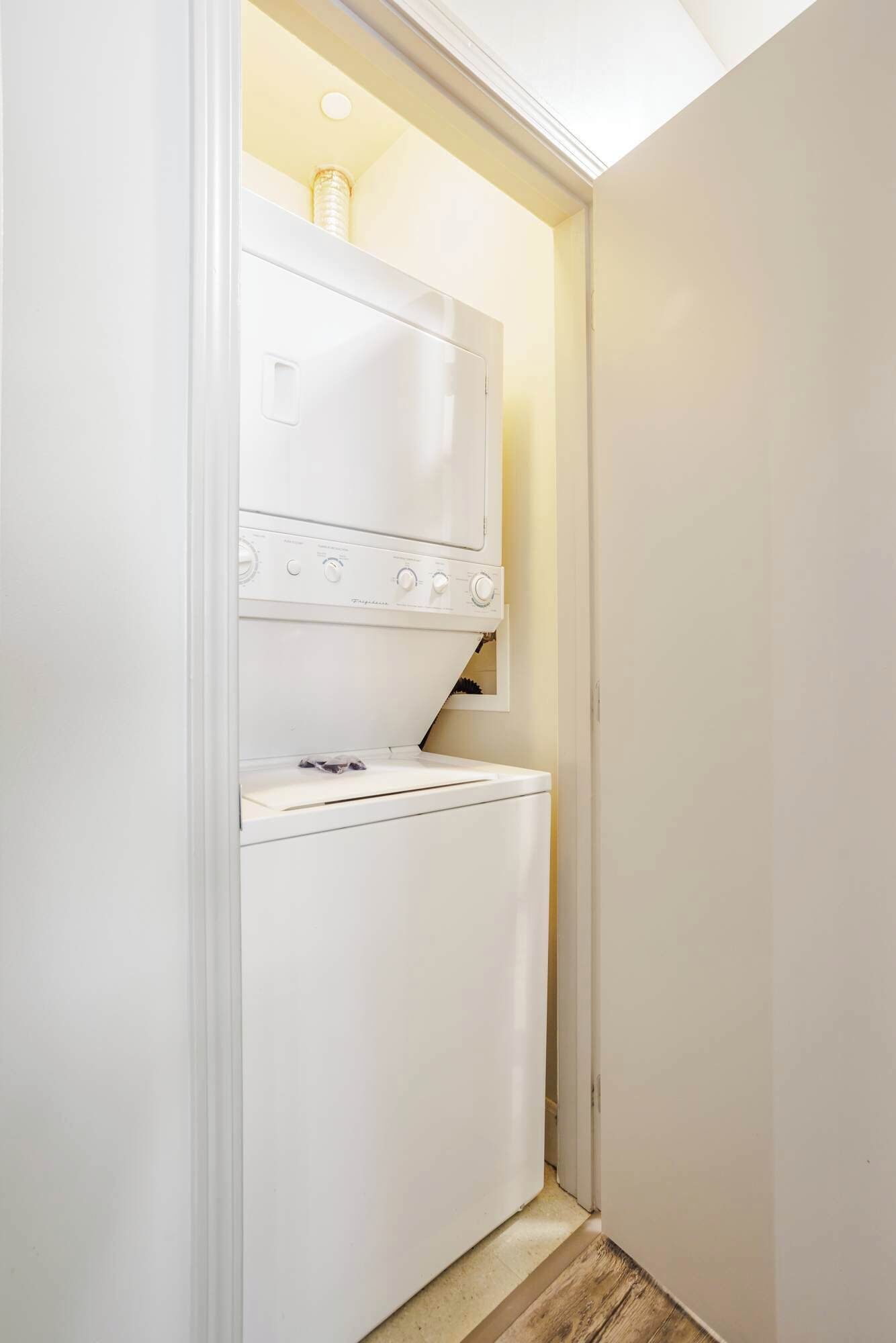 A compact laundry room with a front-loading washing machine and dryer, a small shelf above the dryer, and a wooden floor.