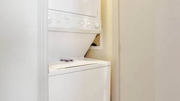 A compact laundry room with a front-loading washing machine and dryer, a small shelf above the dryer, and a wooden floor.