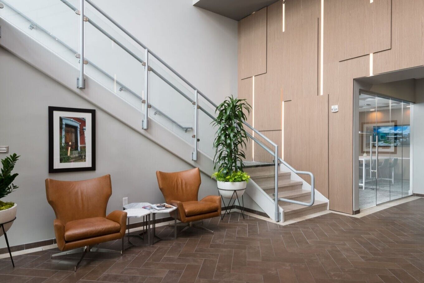 A lobby with a staircase, two leather chairs, a table, a potted plant, and a framed picture on the wall.