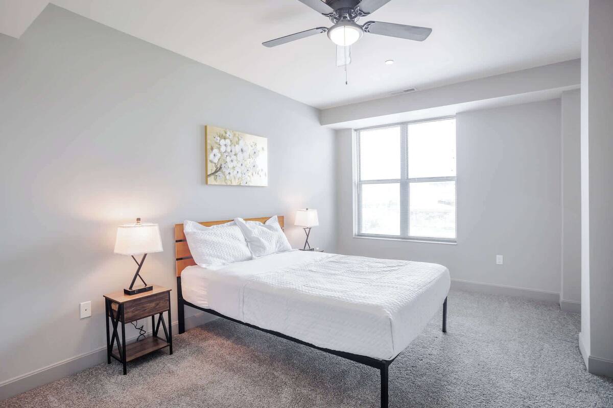 A bedroom with a bed, two bedside tables with lamps, a ceiling fan, a window, and a wall-mounted artwork.