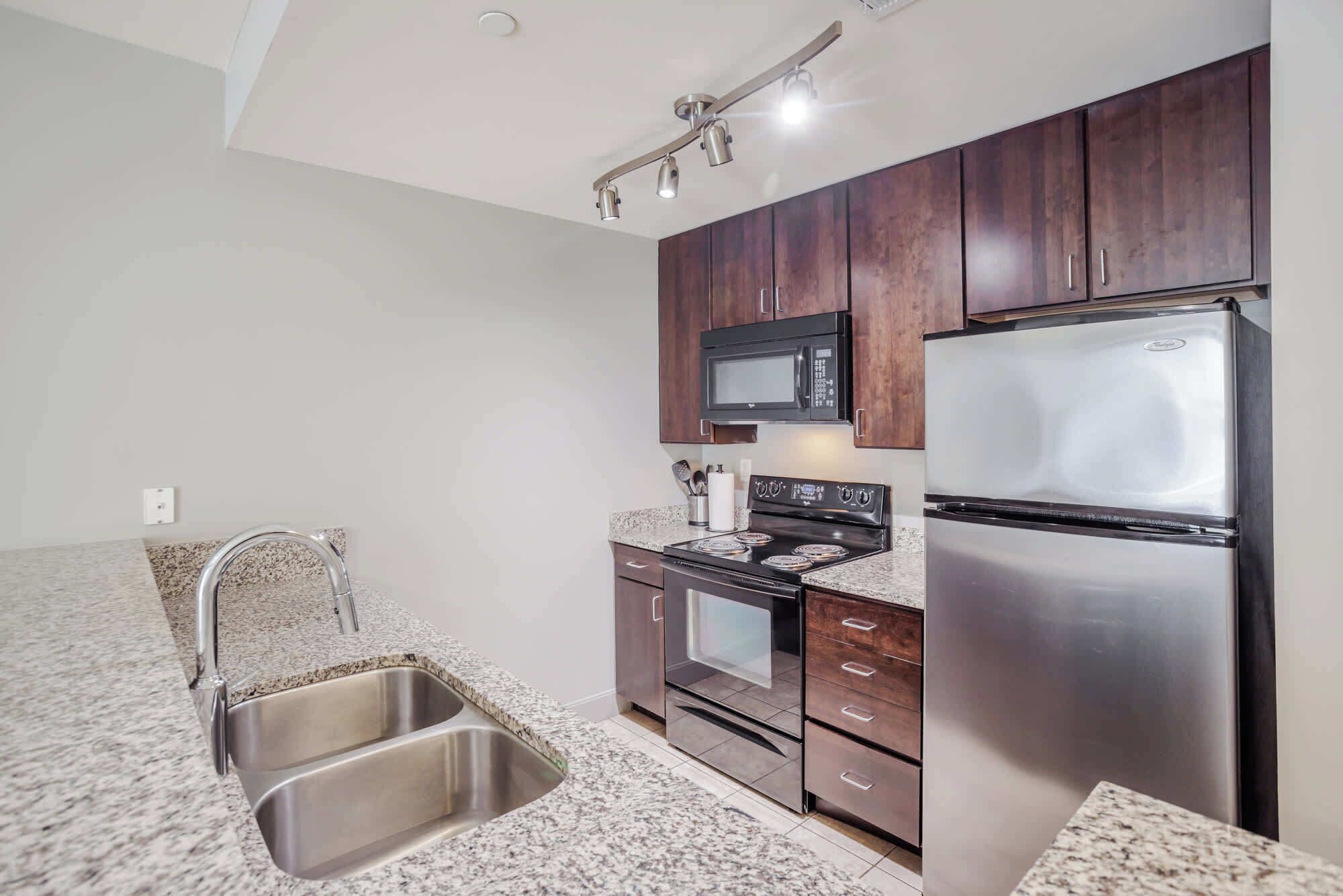 A compact kitchen with dark wooden cabinets, stainless steel appliances including a refrigerator, oven, and microwave, and a granite countertop.