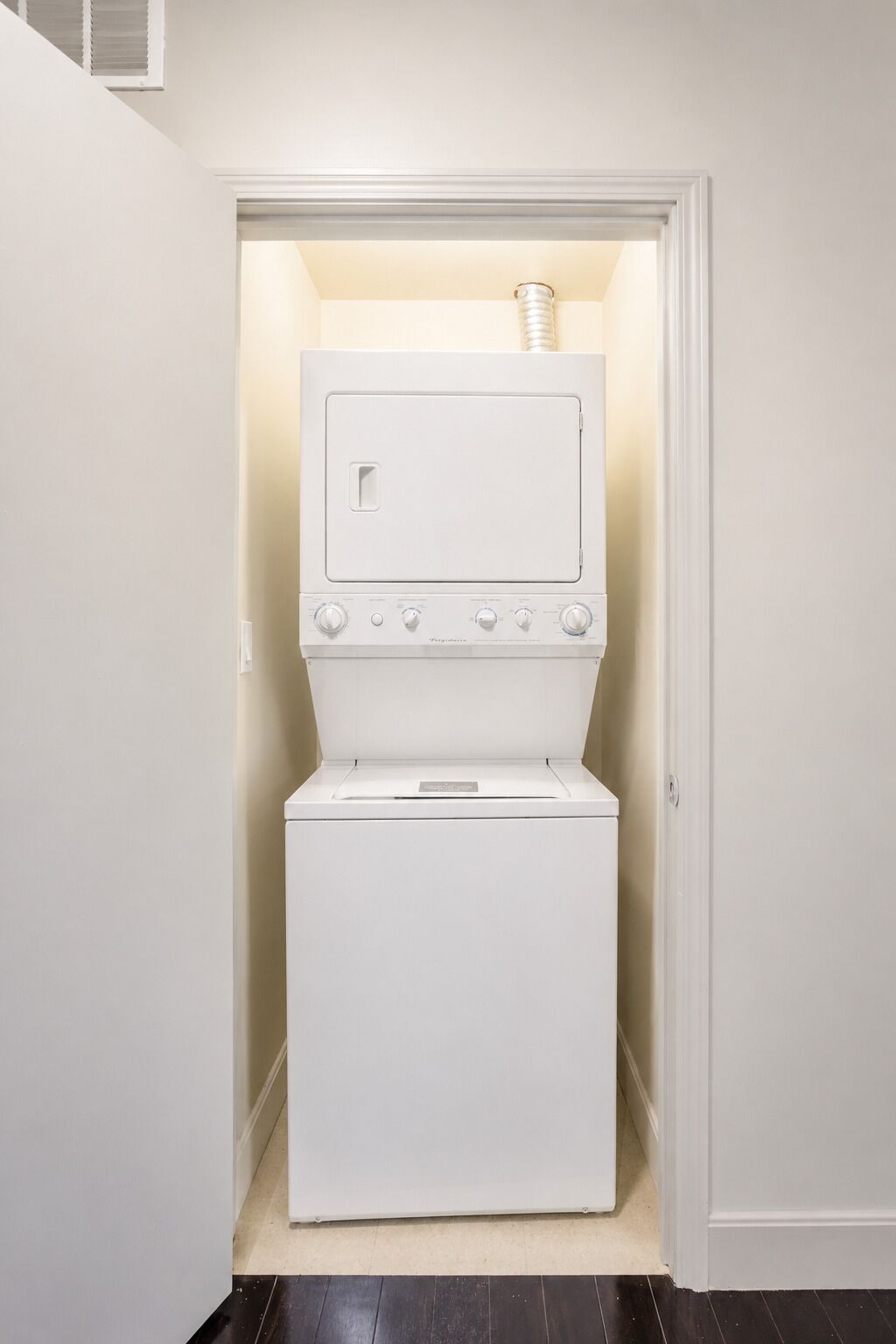 Un lavadero con lavadora y secadora, una rejilla de ventilación en la secadora, y una puerta que da a otra habitación.
