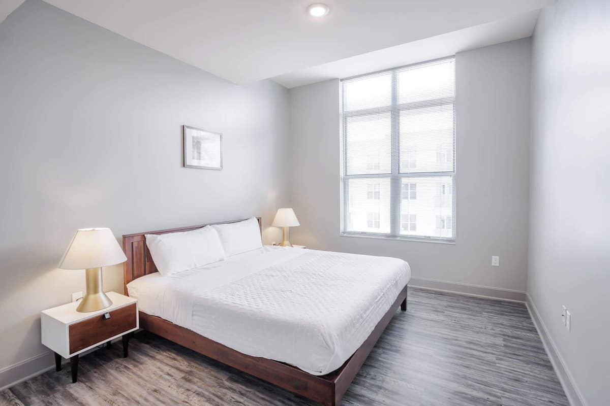 A bedroom with a large bed, two bedside tables with lamps, a picture on the wall, and a window with blinds.