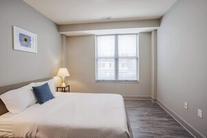 Un dormitorio con una cama, una mesita de noche, una lámpara, una ventana con persianas y un cuadro enmarcado en la pared.