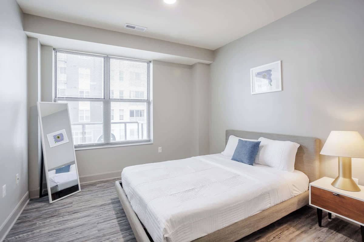 A bedroom with a bed, a bedside table with a lamp, a window with blinds, and a wall-mounted picture.
