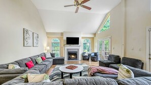 A spacious living room with a fireplace, large windows, and a ceiling fan.