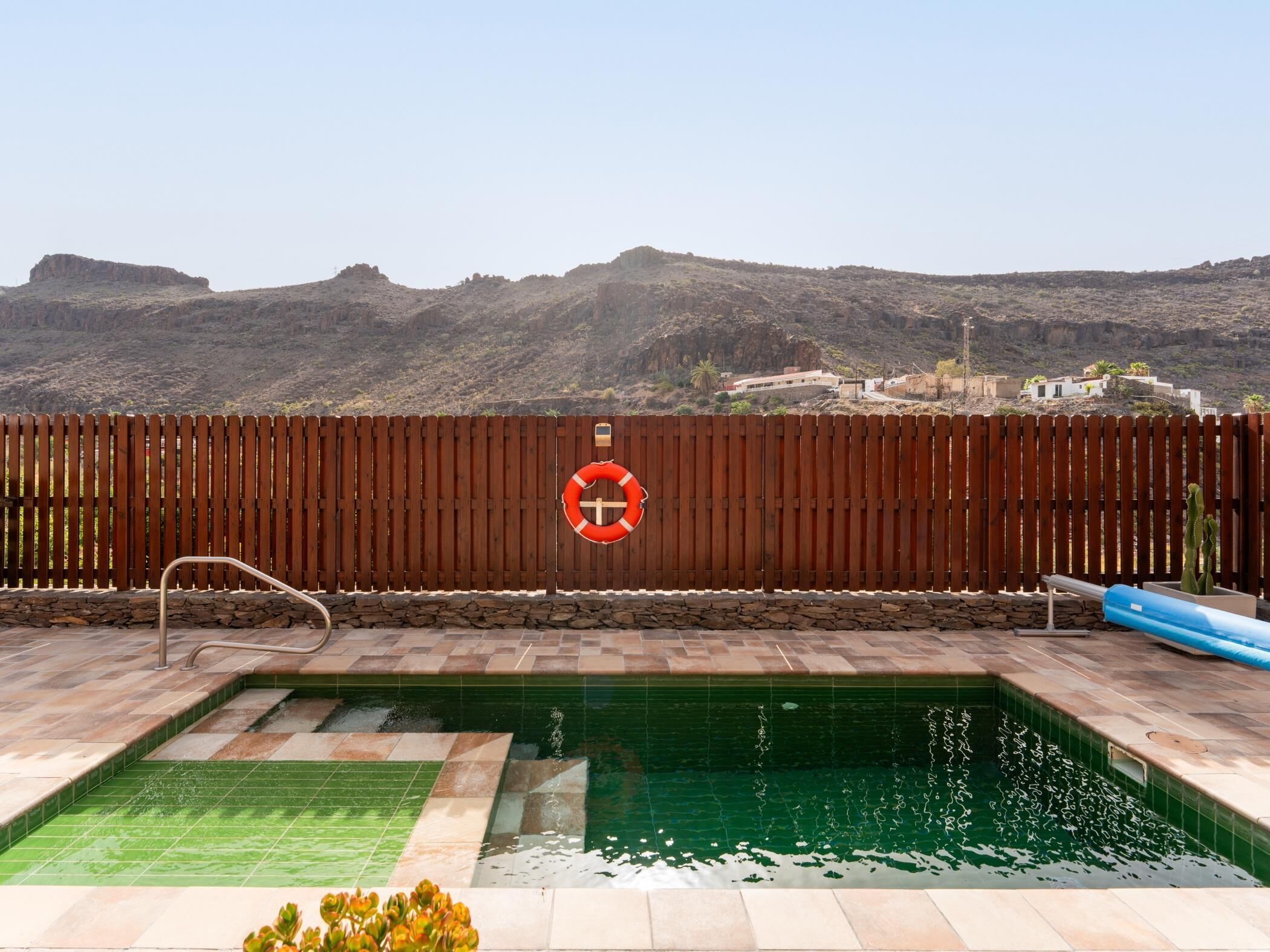 A swimming pool with a wooden fence and a lifebuoy, surrounded by a mountainous landscape.