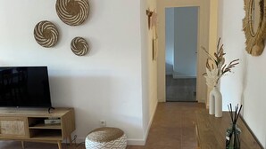 Una sala de estar con televisor, muebles de madera, un otoman tejido y decoración en la pared.