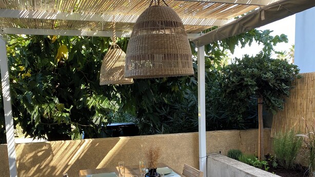Un patio al aire libre con una mesa de madera preparada para cuatro personas, rodeada de sillas y cubierta por un toldo tejido.