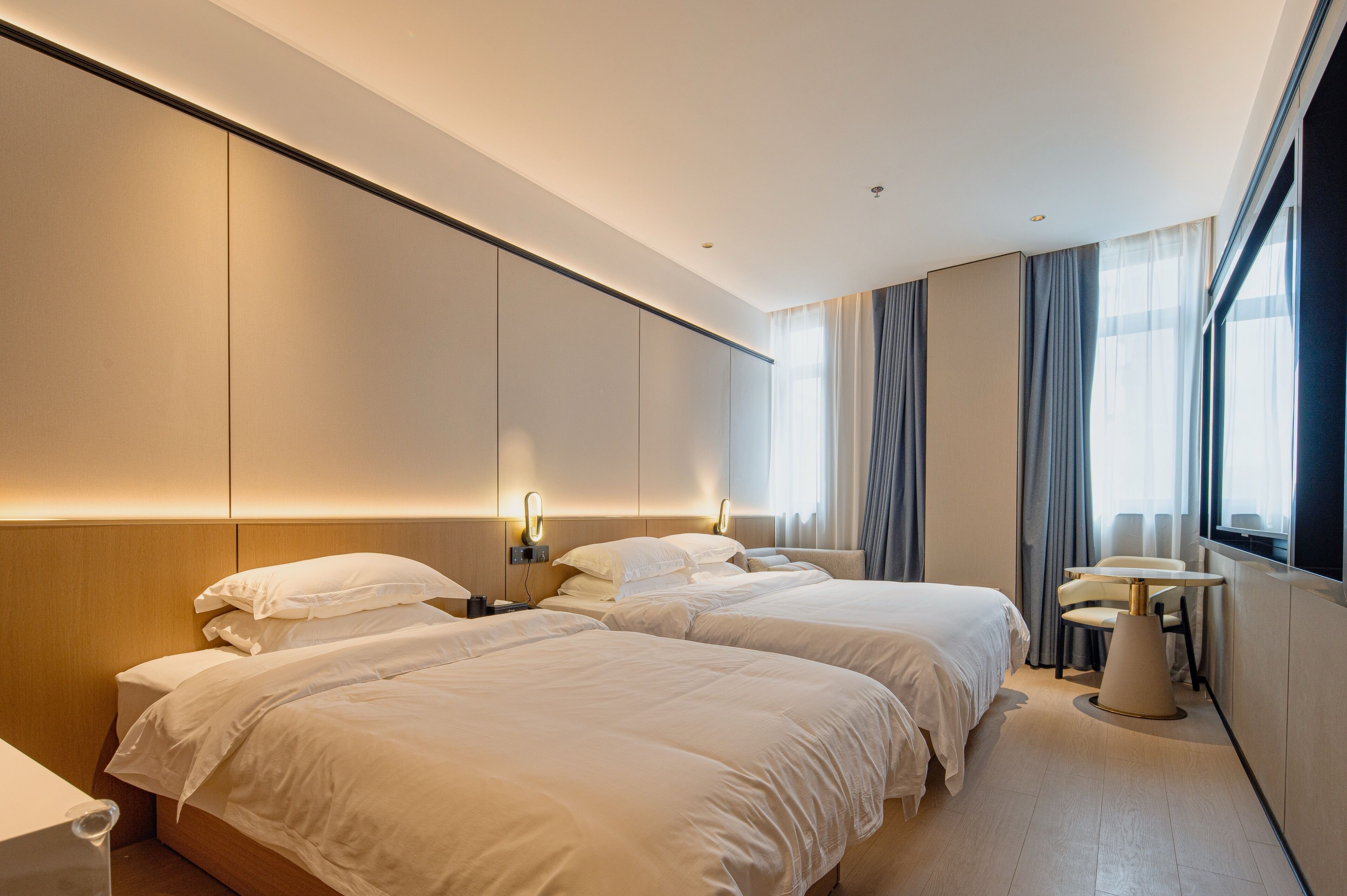 A modern hotel room with two beds, a small table, and a window with curtains.