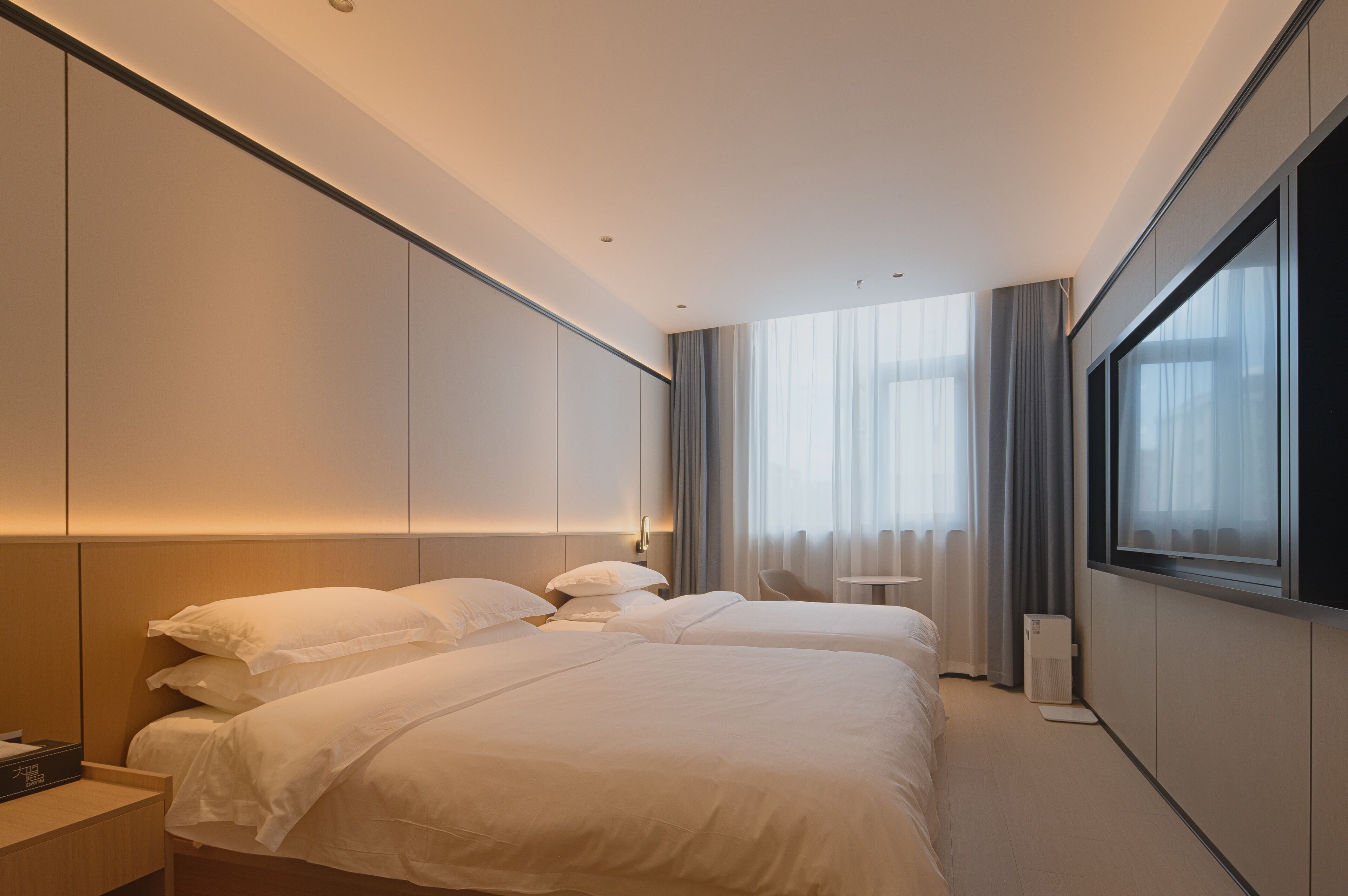 Ein modernes Hotelzimmer mit zwei Betten, einem Nachttisch, einem kleinen Schreibtisch und einem großen Fenster mit Vorhängen.