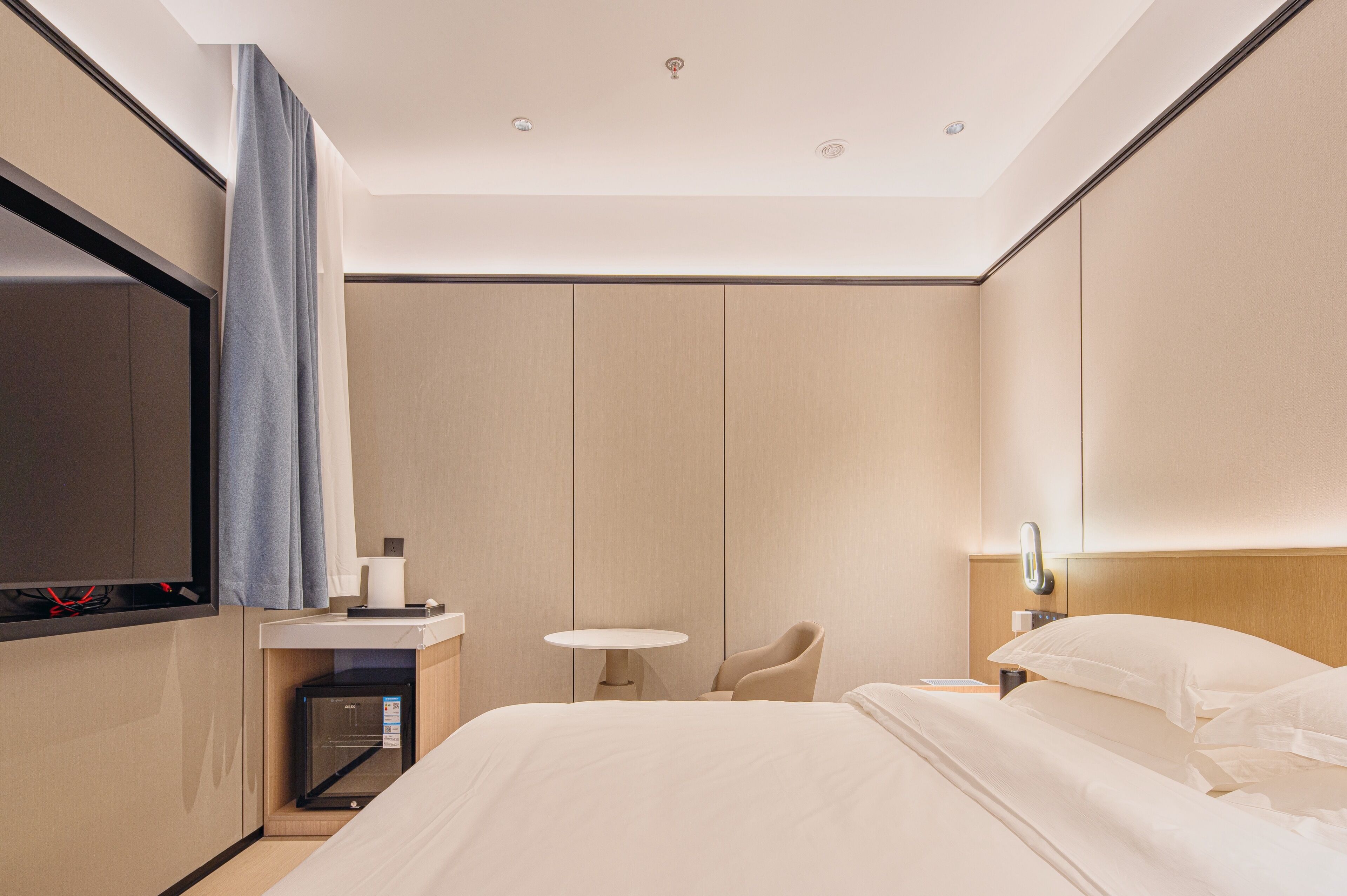 A modern hotel room with a bed, bedside table, chair, and a small desk with a lamp.