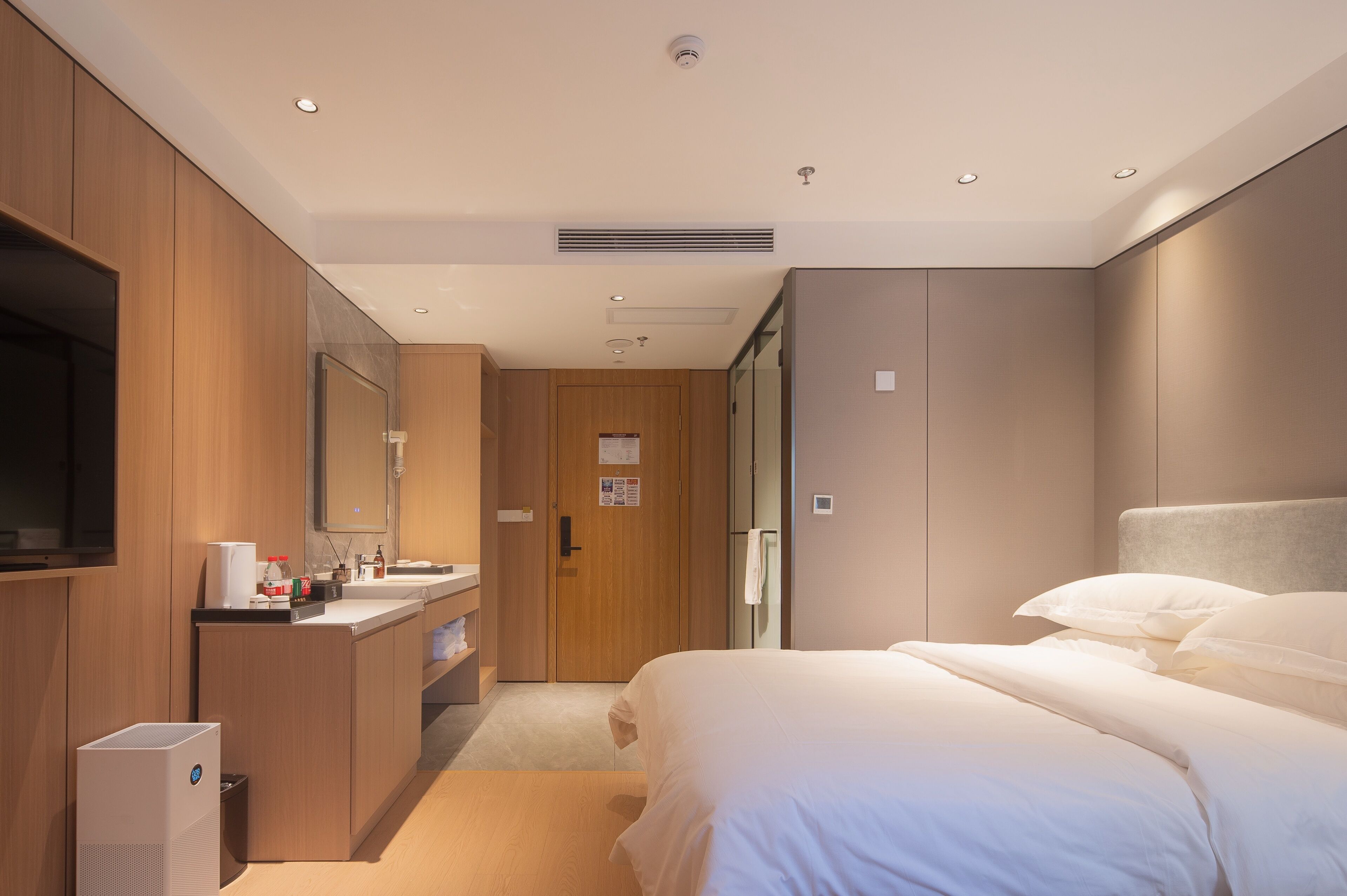 Una habitación de hotel moderna con un armario de madera, televisor integrado, una cama grande con ropa blanca y un baño con lavamanos y espejo.