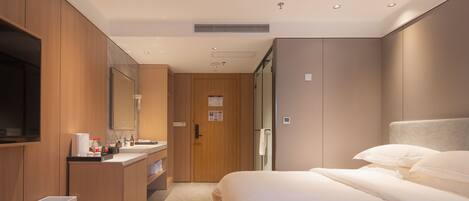 Una habitación de hotel moderna con un armario de madera, televisor integrado, una cama grande con ropa blanca y un baño con lavamanos y espejo.