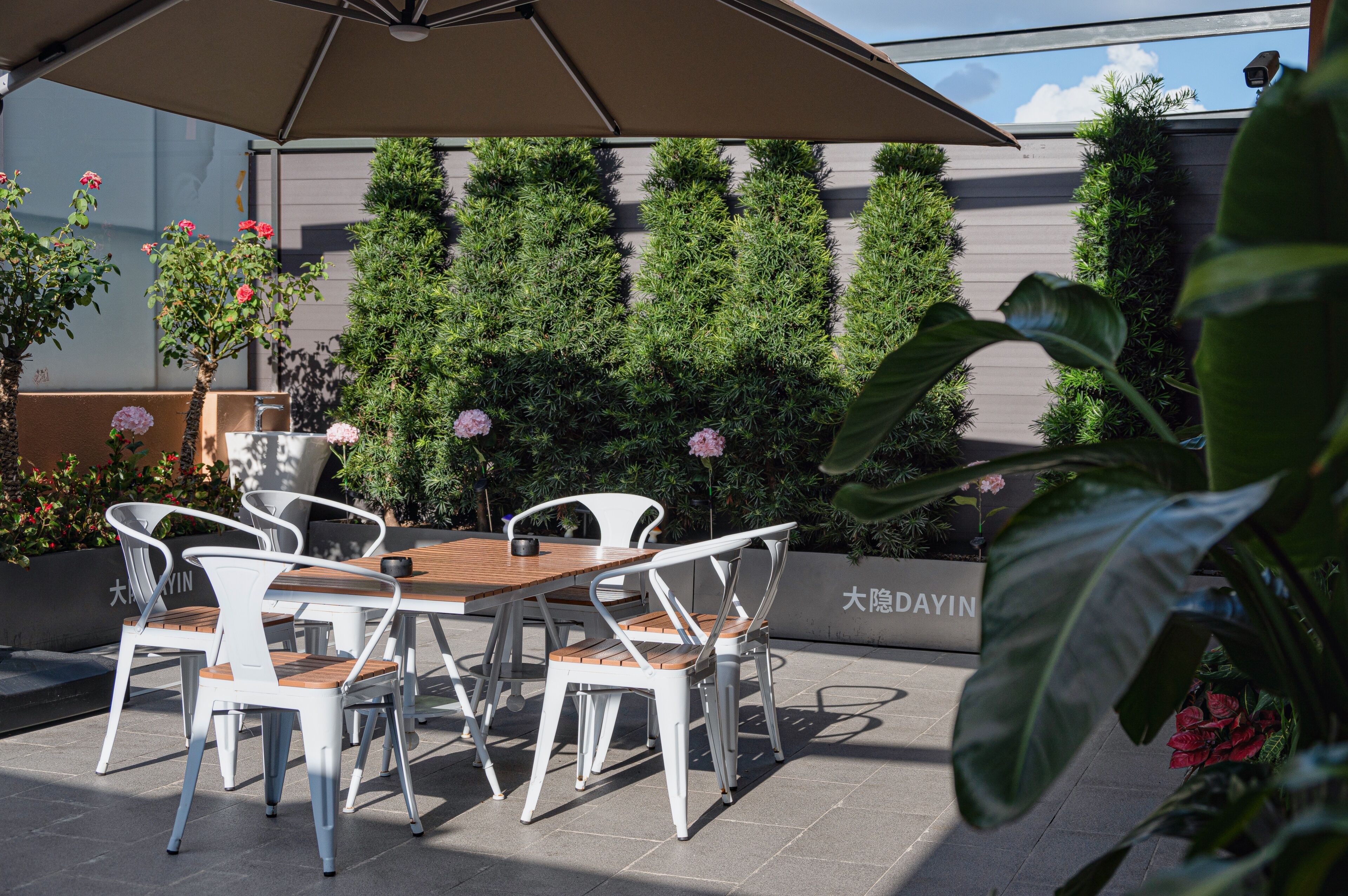 An outdoor patio with a table and chairs, surrounded by neatly trimmed hedges and potted plants.
